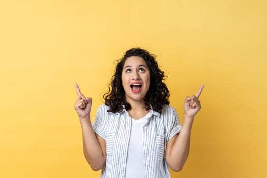 Wow, look, crazy advertisement. Portrait of woman with dark wavy hair pointing up at empty place for ad content and expressing astonishment. Indoor studio shot isolated on yellow background.