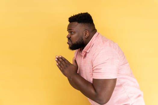 Please, I'm begging forgive. Side view of upset worried man wearing pink shirt looking with imploring desperate grimace, praying for help. Indoor studio shot isolated on yellow background.