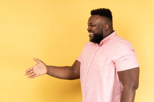 Hello, nice to meet you. Side view of man in pink shirt giving hand to handshake and smiling friendly, consultant or manager offering job, partnership. Indoor studio shot isolated on yellow background