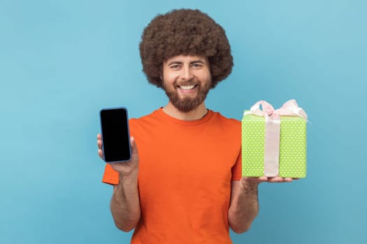 Portrait of satisfied man with Afro hairstyle wearing orange T-shirt standing with present box and showing smart phone with empty display. Indoor studio shot isolated on blue background.