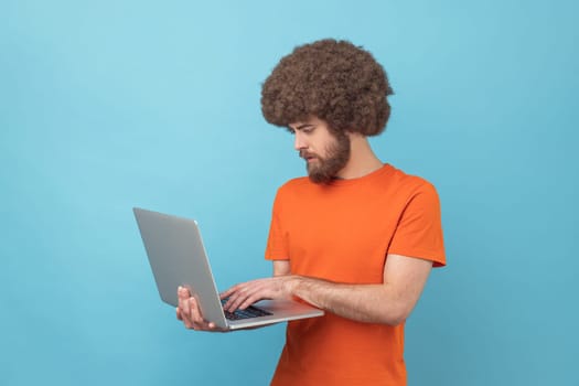 Side view of man with Afro hairstyle wearing orange T-shirt holding laptop in hands, working on computer, looking at display with serious expression. Indoor studio shot isolated on blue background.