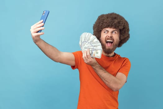 Portrait of excited man with Afro hairstyle wearing orange T-shirt having video call or livestream, showing dollar banknotes to subscribers. Indoor studio shot isolated on blue background.