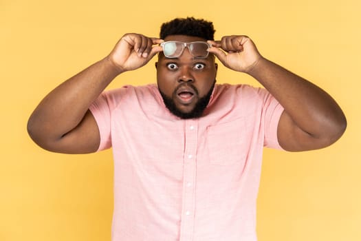 Portrait of shocked amazed surprised man wearing pink shirt standing raised his optical glasses and looking at camera with big eyes and open mouth. Indoor studio shot isolated on yellow background.