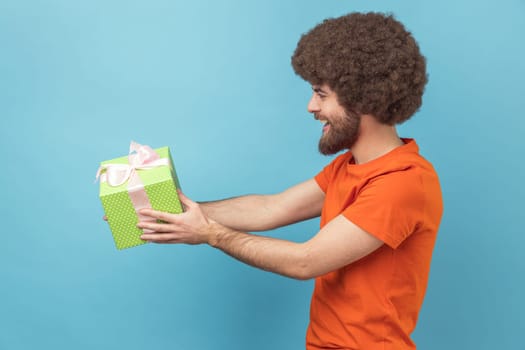 Side view of man with Afro hairstyle wearing orange T-shirt giving gift, congratulating on birthday and offering surprise, holiday bonus wrapped in box. Indoor studio shot isolated on blue background.