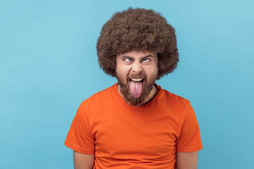 Portrait of man with Afro hairstyle wearing orange T-shirt showing out tongue and crossed eyes with naughty disobedient grimace, making face. Indoor studio shot isolated on blue background.