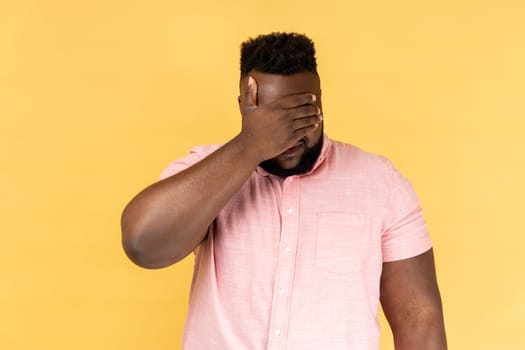 Portrait of man wearing pink shirt closing eyes with hand, dont want to see that, ignoring problems, hiding from stressful situations. Indoor studio shot isolated on yellow background.