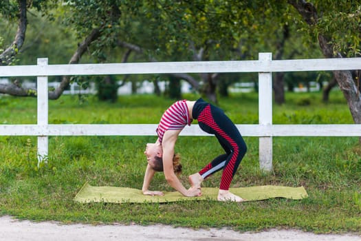 Full length side view of young adult attractive yogi woman practicing yoga, stretching her spine in bridge exercise, urdhva dhanurasana pose, working out outdoor in green park, wearing sportswear.