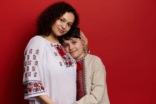 Happy ethnic pregnant woman, expectant mother gently hugging her teen boy, smiling at camera, isolated red background. Mom and son dressed in Ukrainian traditional embroidered shirt. Folklore. Culture