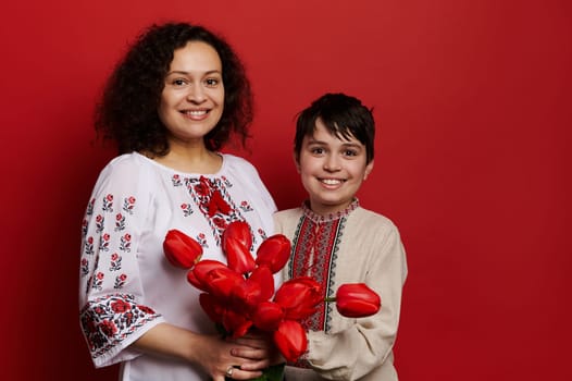 Happy ethnic woman and teen boy, mother and son in traditional Ukrainian embroidered shirts, posing with a bunch of red tulips, smiling looking at camera isolated on red background. Ukraine - culture