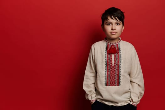 Handsome Caucasian teen boy wearing linen shirt with embroidered ornament in Ukrainian style, looking confidently at camera, isolated on red background. Ukrainian people, culture and traditions