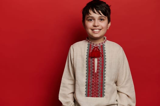 Portrait of smiling Caucasian teen boy in Ukrainian traditional embroidered linen shirt, isolated on red backdrop. Ukrainian people, culture, folklore, traditions. Vyshyvanka - Eastern Slavic fashion