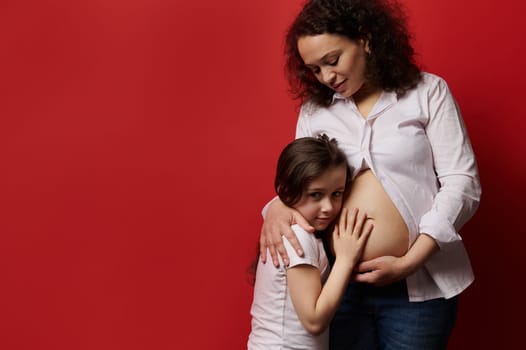 Multi ethnic middle aged gravid beautiful woman, pregnant expectant mother with naked belly gently hugging her lovely daughter, standing isolated on red background. Love. Pregnancy. Family. Maternity