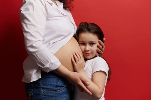 Beautiful little girl hugging the belly of her pregnant mother, looking at camera, isolated on red background. Gravid woman with naked tummy embracing her lovely daughter. Pregnancy. Family. Maternity