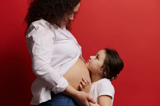 Caucasian happy little child girl hugging the belly of her pregnant mom, isolated on red color background. Happy Mother's Day. Pregnancy. Motherhood. Maternity leave. Family values and relationships