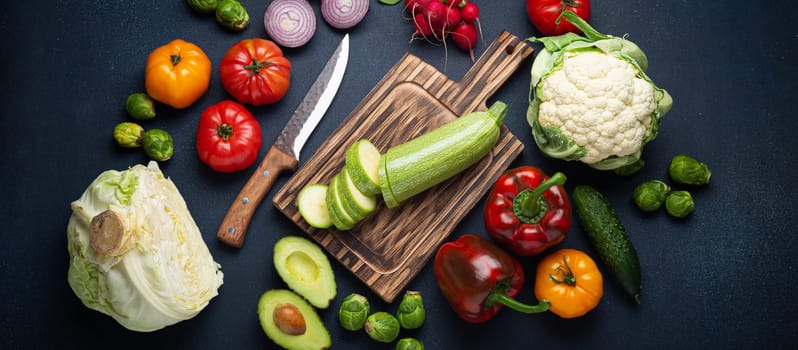 Fresh various vegetables, cut zucchini on wooden cutting board and knife on rustic dark background top view. Cooking vegetarian meal from healthy ingredients, diet food and nutrition concept