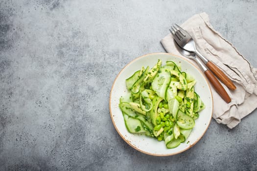 Healthy vegan green avocado salad bowl with sliced cucumbers, edamame beans, olive oil and herbs on ceramic plate top view on grey stone rustic table background. Space for text