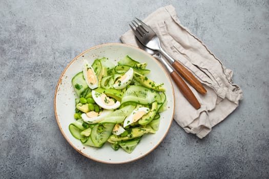 Healthy green avocado salad bowl with boiled eggs, sliced cucumbers, edamame beans, olive oil and herbs on ceramic plate top view on grey stone rustic table background