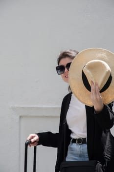 young happy brunette girl in sunglasses and straw hat with a suitcase goes on vacation, a woman stands against the background of a white brick wall, High quality photo