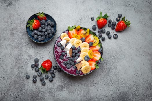 Healthy summer acai smoothie bowl with chia seeds, fresh banana, strawberry, blueberry, cocos, kiwi top view on rustic concrete background with spoon