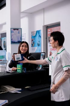 Apothecary customer asking pharmacist about vitamin at checkout, showing medical product package. Young asian woman buying food supplements and discussing medication with pharmacy cashier