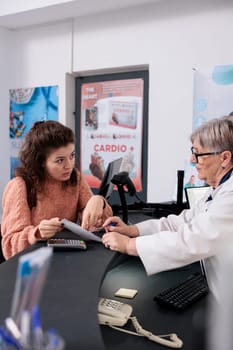 Elderly pharmacist standing at drugstore counter helping customer with pharmaceutics treatment after checking medical prescription, discussing pills leaflet. Health care service and support