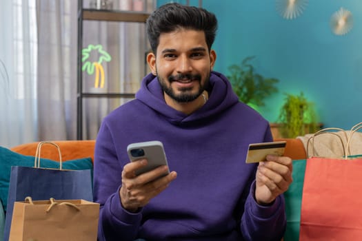 Happy shopaholic consumer indian man sitting with shopping bags at modern home apartment making online order, payment with credit plastic bank card. Discount sale on internet. Young hindu guy on sofa