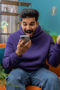 Happy indian man use mobile smartphone typing browsing say Wow yes found out great big win good news celebrate lottery jackpot doing winner gesture. Hindu young guy at home in room sitting on sofa