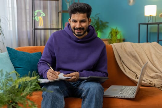 Indian man freelancer work with documents on home couch, reading information, business papers prepare financial report on laptop. Young hindu guy accountant analyzing graph documents, calculate taxes