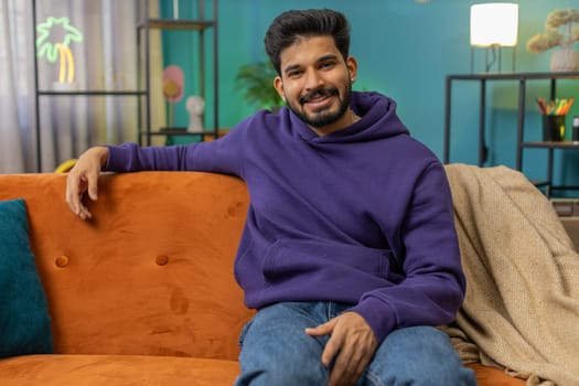 Portrait of happy calm indian man at home couch smiling friendly, glad expression looking away dreaming resting, relaxation feel satisfied concept good news, celebrate win. Hindu guy in living room