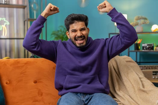 Happy indian man shouting, celebrating success, winning, goal achievement good news raising fists in gesture I did it, lottery luck at modern home apartment. Hindu guy in living room sitting on couch