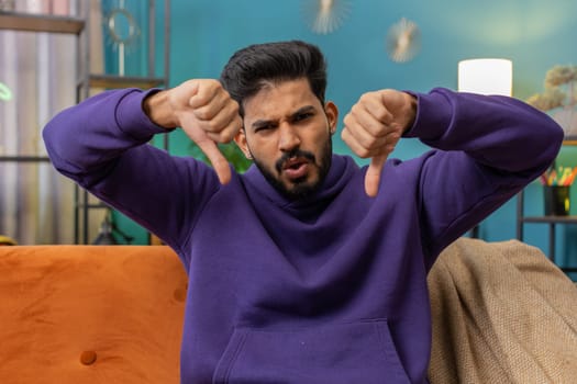 Dislike. Upset indian man showing thumbs down sign gesture, expressing discontent, disapproval dissatisfied bad work at modern home apartment indoors. Displeased hindu guy in living room on sofa
