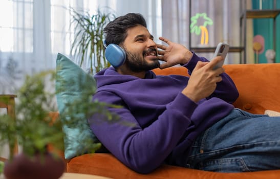 Happy overjoyed hindu indian man in wireless headphones relaxing sits on couch at home apartment choosing listening favorite energetic disco dancing music. People weekend daytime leisure activities