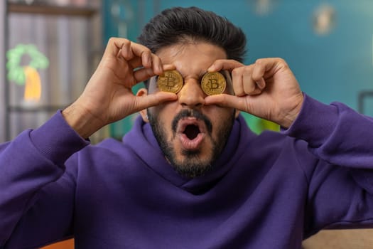 Indian man freelancer holding two gold coins successful developer programmer. Hindu guy stock trader earning bitcoins after online monitoring trading operations. Increasing wealth financial prosperity