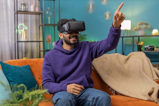 Indian man using virtual reality futuristic technology VR app headset helmet to play simulation 3D 360 video game, watching film movie at modern home apartment. Hindu guy in goggles sitting on couch