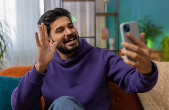 Happy indian man blogger taking selfie on smartphone, communicating video call online with social media subscribers followers, recording vlog stories. Young hindu guy at home apartment room on couch