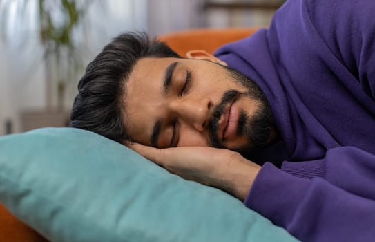 Tired indian man lying down in bed taking a rest. Carefree young hindu guy napping, falling asleep on comfortable sofa with pillows at home apartment room. Closed his eyes enjoy daytime nap alone