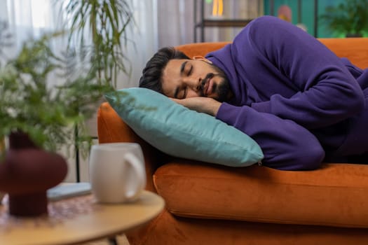Tired indian man lying down in bed taking a rest. Carefree young hindu guy napping, falling asleep on comfortable sofa with pillows at home apartment room. Closed his eyes enjoy daytime nap alone