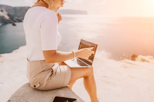 Successful business woman in yellow hat working on laptop by the sea. Pretty lady typing on computer at summer day outdoors. Freelance, travel and holidays concept.