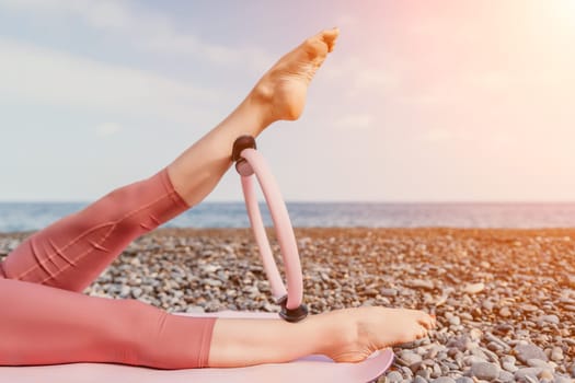 Middle aged well looking woman with black hair doing Pilates with the ring on the yoga mat near the sea on the pebble beach. Female fitness yoga concept. Healthy lifestyle, harmony and meditation.