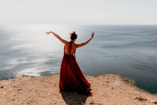 Side view a Young beautiful sensual woman in a red long dress posing on a rock high above the sea during sunrise. Girl on the nature on blue sky background. Fashion photo.