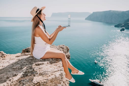 Successful business woman in yellow hat working on laptop by the sea. Pretty lady typing on computer at summer day outdoors. Freelance, travel and holidays concept.