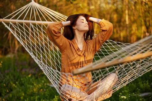 a happy woman in a long orange dress is resting lying in a hammock at the dacha, straightening her long hair and looking away, illuminated by the summer sun during sunset. High quality photo