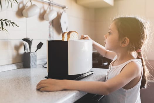 5 - 7 year old kid girl who is preparing breakfast from toast in a toaster in cozy home interior, selective soft focus.