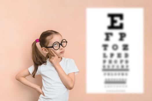 Thoughtful smart girl of elementary school age in diopter glasses against the background of a blurred snellen table for checking visual acuity.