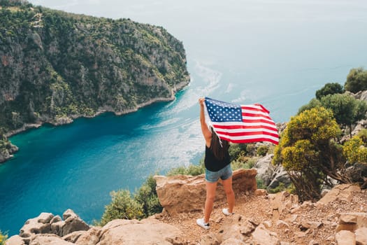 Young woman standing on a rock cliff and waving the US flag while looking at sea beneath. Girl traveller waving the American flag while standing on a mountain top. 4 fourth July Independence Day