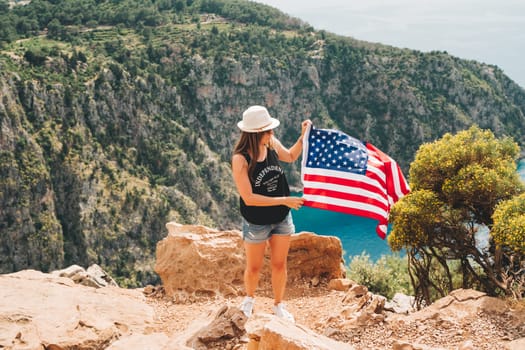 Young woman standing on a rock cliff and waving the US flag while looking at sea beneath. Girl traveller waving the American flag while standing on a mountain top. 4 fourth of July Independence day