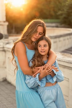 Portrait of mother and daughter in blue dresses with flowing long hair against the backdrop of sunset. The woman hugs and presses the girl to her. They are looking at the camera