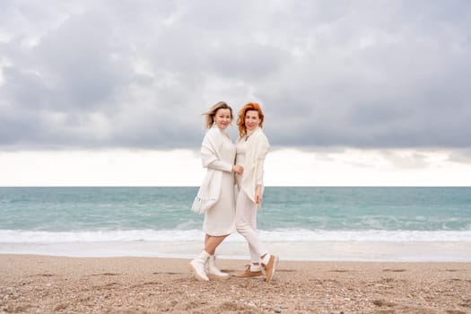 Women sea walk friendship spring. Two girlfriends, redhead and blonde, middle-aged walk along the sandy beach of the sea, dressed in white clothes. Against the backdrop of a cloudy sky and the winter sea. Weekend concept