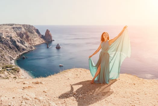 Side view a Young beautiful sensual woman in a red long dress posing on a rock high above the sea during sunrise. Girl on the nature on blue sky background. Fashion photo.