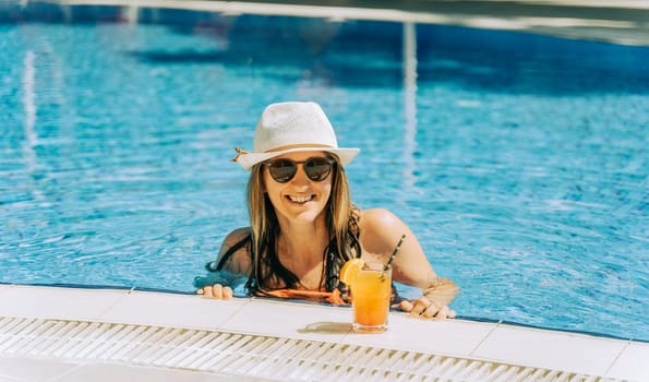 Portrait of beautiful cute smiling young woman lady girl in a bikini, hat, and sunglasses holding tropical lemonade cocktail while chilling in swimming pool. Hello summer holiday vacation